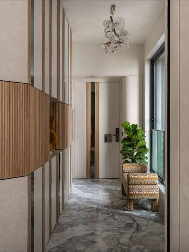 Marble-floored foyer corridor with fluted wood panelling, glass chandelier, and plant beside a woven stool