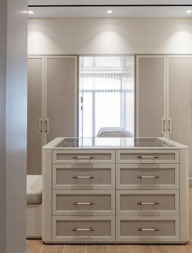 Beige walk-in closet with central glass-top drawer island and mirrored wardrobe doors reflecting bedroom window