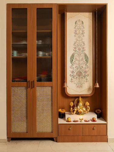 Wooden pooja unit with cane doors beside a niche housing deity idols and a peacock-motif scroll
