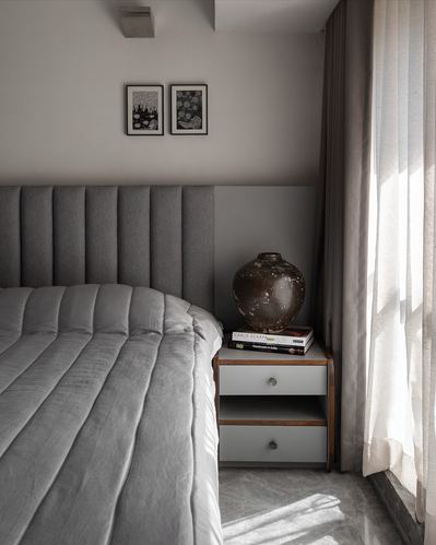 Grey channel-tufted bed beside nightstand with ceramic vase and sheer curtain light