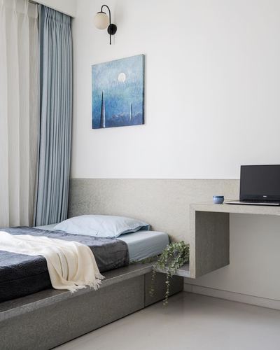 Minimalist bedroom with grey stone platform bed, blue curtains, and floating desk with laptop.