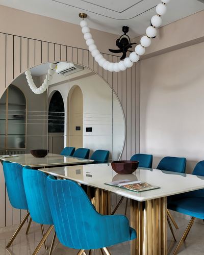 Blush dining room with teal velvet chairs, marble-topped brass table, and arched mirror reflecting a bubble chandelier