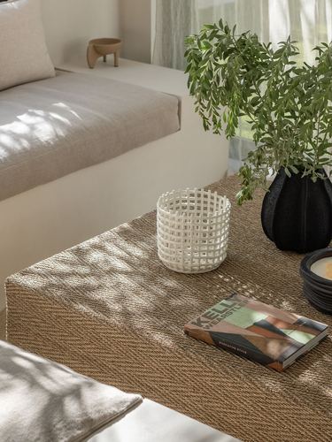 Woven coffee table with ceramic vases, book, and candle under dappled sunlight, sofa behind