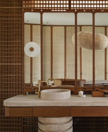 Travertine vessel sink and brass faucet on teak vanity beneath a cane-paneled mirror wall