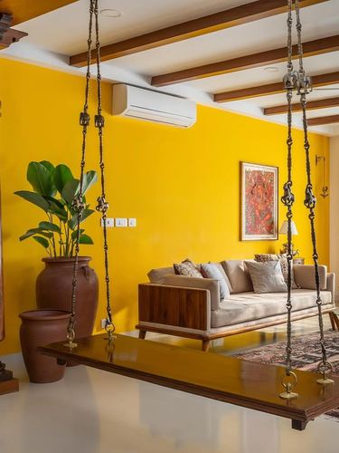 Mustard yellow living room with wooden ceiling beams, hanging jhula, terracotta pots, beige sofa, and framed deity artwork