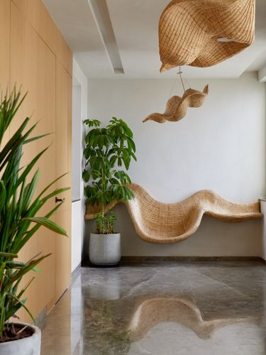Wicker sculptural bench along a wall beneath a matching hanging wicker pendant, near a wood-paneled door and plants