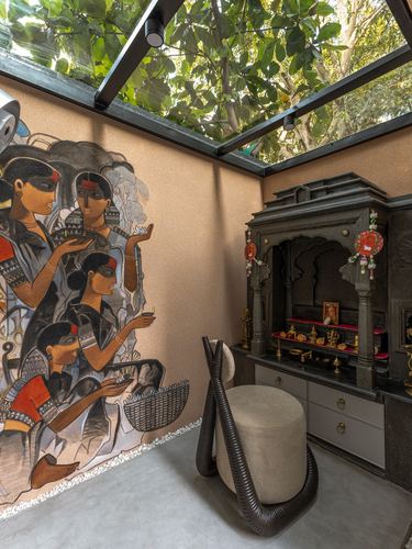 Carved stone pooja unit beside a figurative mural under a glass skylight roof with tree canopy above