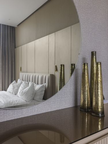 Round mirror reflecting a tufted bed and fluted wardrobe beside tall bronze glass vases