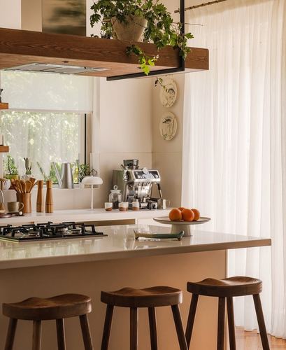Kitchen island with wooden stools, gas cooktop, and espresso station under sunlit sheer curtains