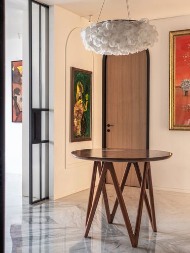 Marble-floored foyer with round wooden table, arched wood door, and framed paintings under a petal-shaped pendant