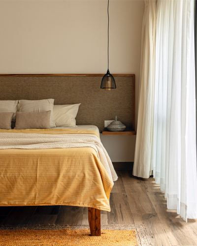 Bed with mustard throw beside woven headboard, pendant light, and sheer curtains on wood floor