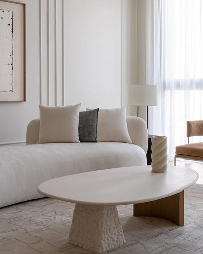 Minimalist living room with curved beige sofa, oval plaster coffee table, and sheer curtains.