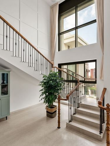Double-height foyer with wooden-railed staircase, large windows, and a potted plant beside a mint cabinet