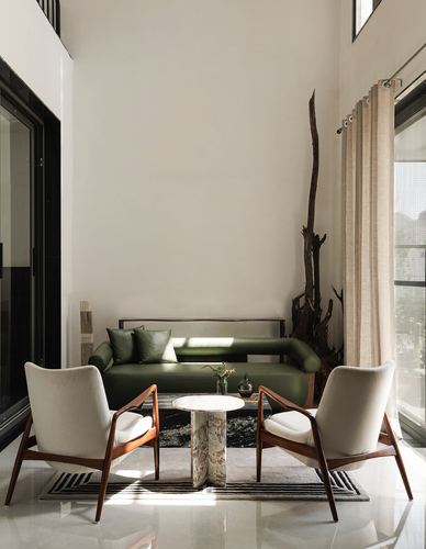 Double-height living room with green leather sofa, two teak armchairs, and a driftwood sculpture beside curtained glass doors