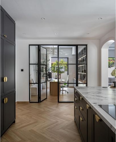 Dark kitchen island with marble top opens through glass doors to a study with herringbone flooring.