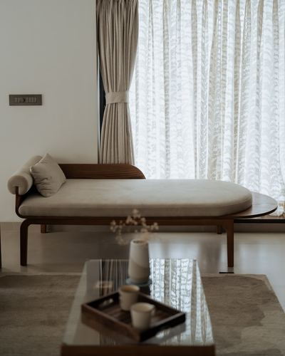 Curved wooden daybed with cream cushions beside sheer curtained window in minimalist living room