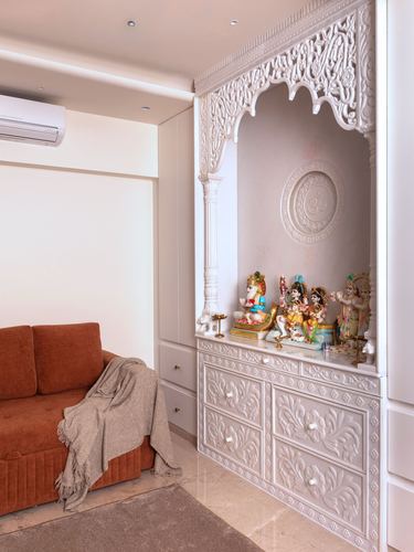 White carved pooja cabinet with arched niche and deity idols beside a terracotta sofa
