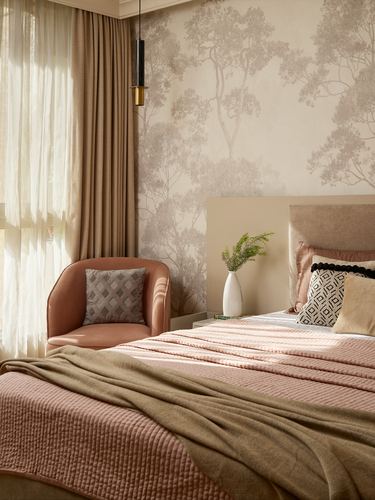 Bedroom with tree-print wallpaper, pink armchair, and layered pink and olive bedding in soft light
