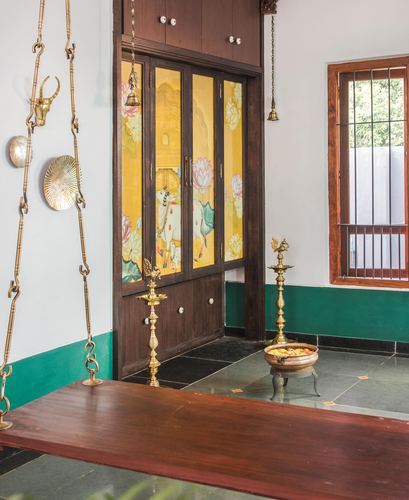 Traditional pooja alcove with painted glass cabinet, brass lamps, and hanging wooden swing in Pondicherry home