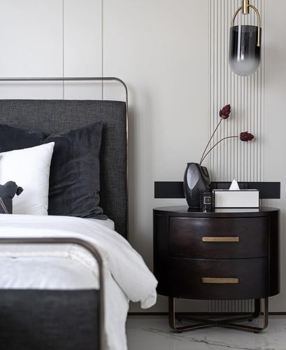 Dark curved nightstand beside upholstered bed with brass pendant and dried flowers in vase