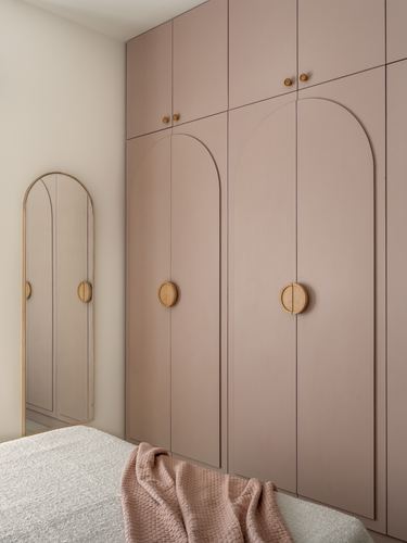 Dusty pink arched wardrobe with round wooden handles beside an arched mirror in bedroom corner