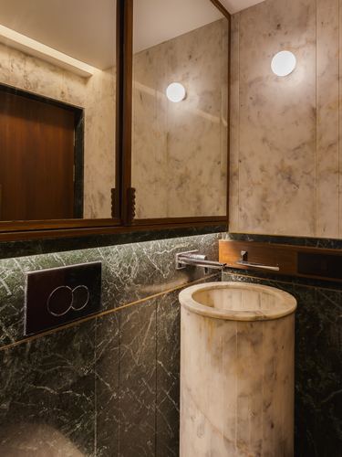 Bathroom with cylindrical marble pedestal sink, dark green stone wall, wood-framed mirror cabinet, round sconce light