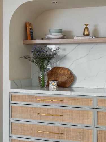 Kitchen alcove with marble backsplash, wood shelf styled with plates, and cane-front drawers with brass handles