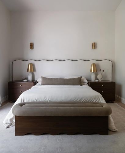 Symmetrical bedroom with wavy headboard, brass sconces, dark wood nightstands, and upholstered scalloped bench.