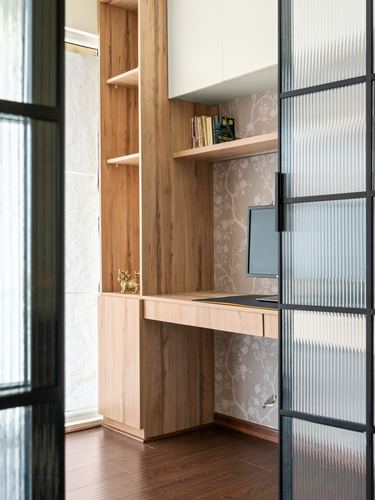 Wood-paneled study nook with built-in desk, monitor, and floral wallpaper seen through a fluted glass door.