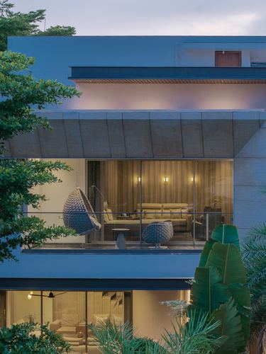 Twilight facade of concrete villa with glowing glass balcony, hanging cane chair, and lush foliage