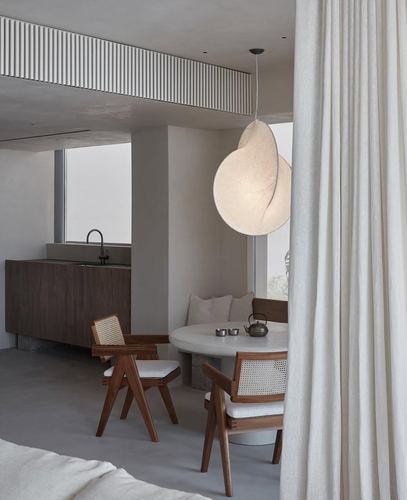 Minimalist dining nook with cane chairs, round stone table, and sculptural pendant light beside sheer curtain