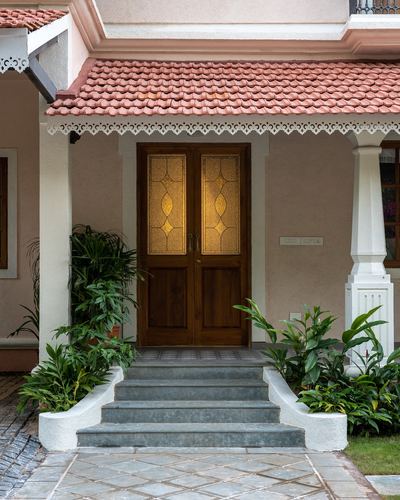 Goan villa entrance with terracotta tile roof, glowing stained-glass wooden door, and stone steps flanked by plants