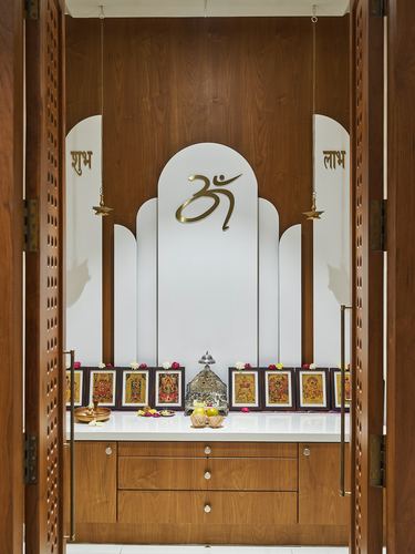 Pooja room with arched white niche, om symbol, deity frames, and hanging brass diyas flanked by carved wooden doors