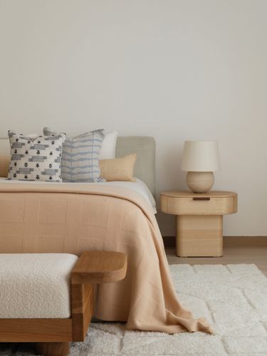 Bedroom corner with peach bedspread, patterned pillows, curved wood nightstand and lamp, boucle bench.