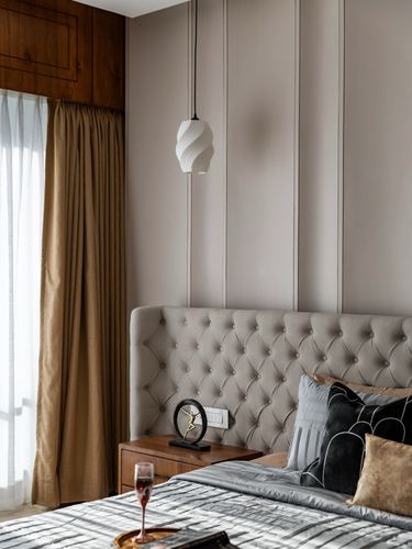 Tufted grey headboard beside wood bedside table under a sculptural pendant light in a warm-toned bedroom.