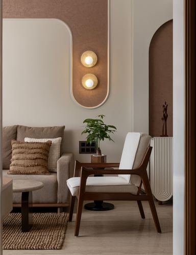 Curved beige-and-terracotta wall niche with brass sconces above a wooden accent chair and sofa