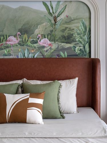 Bed with rust leather headboard beneath an arched flamingo mural, styled with green and cream pillows
