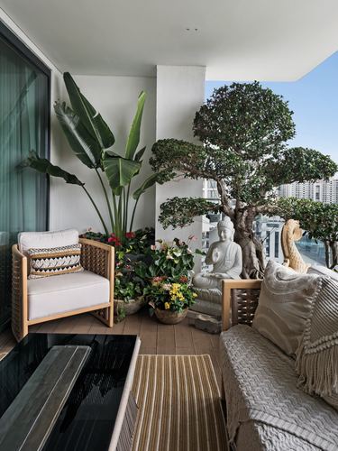 Balcony with rattan armchair, sofa, potted plants, bonsai tree and stone buddha statue overlooking city skyline