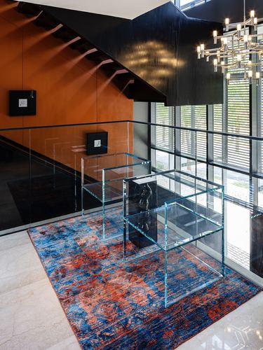 Double-height stairwell landing with orange wall cladding, glass furniture, and a blue-orange rug
