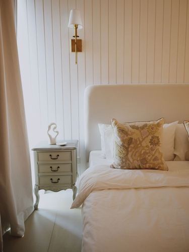 Cream upholstered bed with embroidered pillow beside a vintage-style nightstand and brass sconce