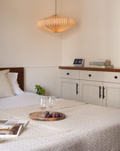 Bedroom corner with woven pendant light, bed with tray of wine glasses, and cabinet