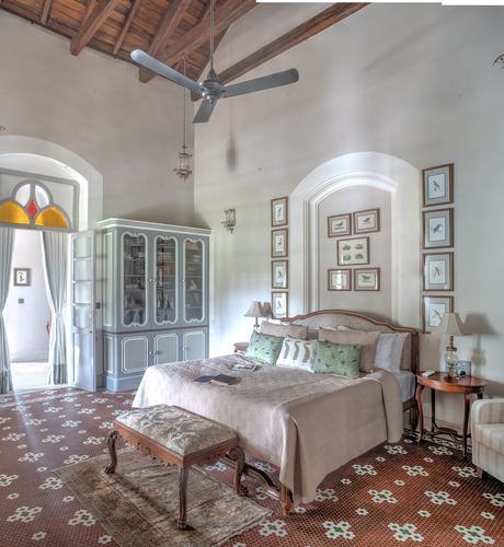 High-ceilinged heritage bedroom with patterned tile floor, arched niche of framed bird prints, and carved wooden bed