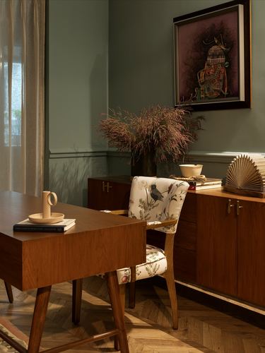 Study nook with wooden desk, printed armchair, sideboard, and framed folk art on sage wall