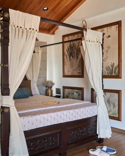 Carved dark wood four-poster bed with cream curtains under a sloped wooden ceiling, framed botanical prints on wall