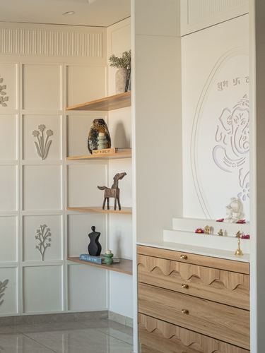 White panelled corner shelving beside a pooja unit with wave-carved wood drawers and an oval elephant motif niche