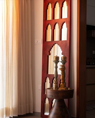 Red carved arched wooden panel with brass candlesticks on a round wooden table beside sheer curtains