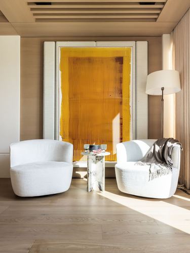 Two white boucle chairs flank a marble side table beneath a large mustard artwork in a sunlit room
