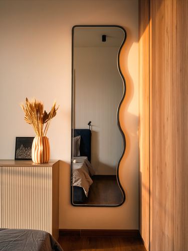 Wavy black-framed mirror reflecting a bedroom, beside a fluted cabinet with dried flower vase