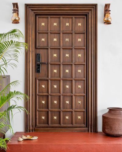 Wooden paneled front door with brass studs flanked by lantern sconces, terracotta pot and plant beside it