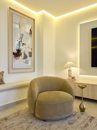 Curved velvet armchair in warm-lit living room corner with abstract art and cove lighting